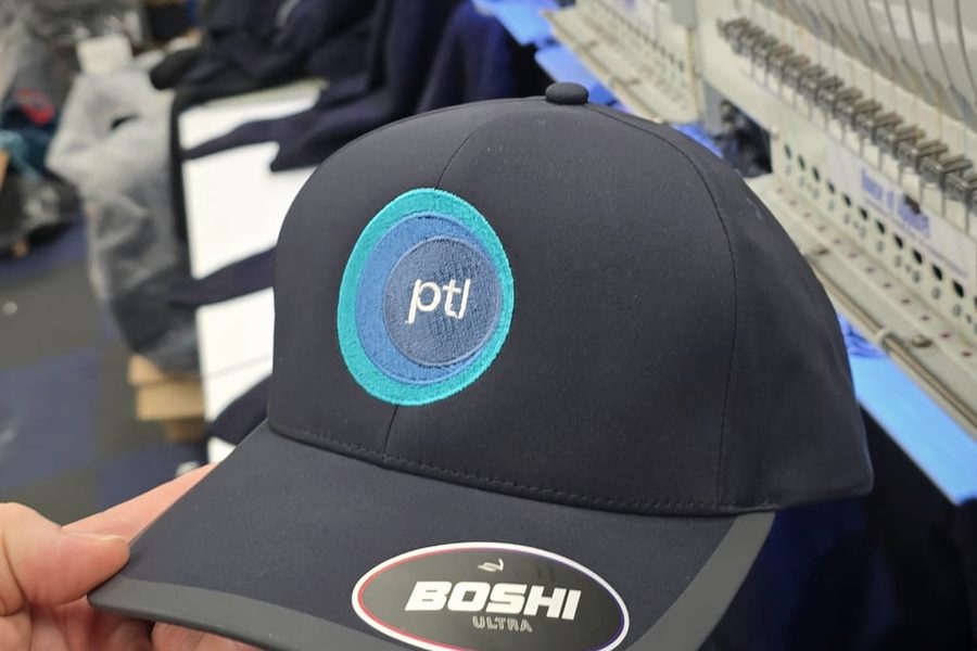 A navy cap with an embroidered PTL logo, in front of multi-head embroidery machines